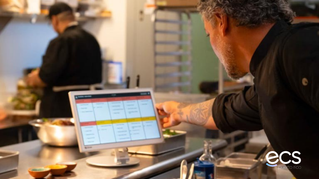 Chef en una cocina profesional usando una pantalla del sistema KDS para revisar y organizar pedidos digitales mientras otro cocinero trabaja al fondo. La imagen ilustra la gestión de pedidos en tiempo real en cocina con tecnología de ECS Puerto Rico.